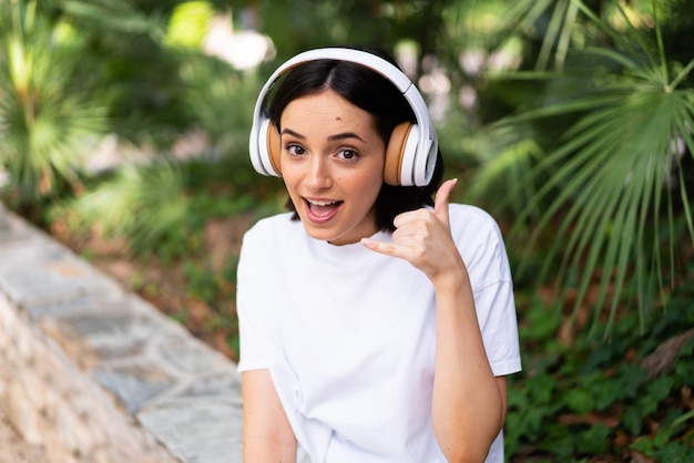 Jeune femme caucasienne avec des écouteurs à l'extérieur faisant le geste du téléphone. Rappelez-moi signe