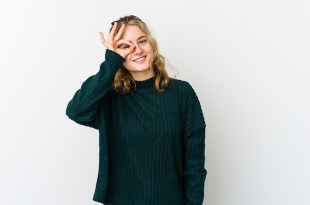 Jeune femme caucasienne sur fond blanc excité en gardant le geste ok sur les yeux.