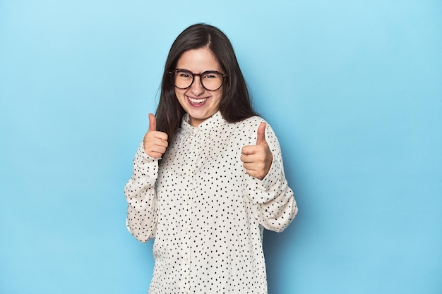 Jeune femme caucasienne sur fond bleu levant les deux pouces vers le haut souriante et confiante