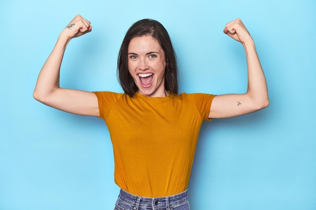 Jeune femme caucasienne sur fond bleu montrant un geste de force avec les bras symbole du pouvoir féminin