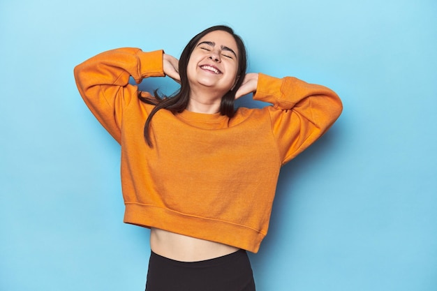 Jeune femme caucasienne sur fond bleu se sentant en confiance avec les mains derrière la tête