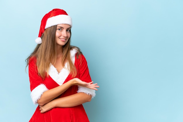 Jeune femme caucasienne habillée en maman noel isolée sur fond bleu présentant une idée tout en regardant en souriant vers