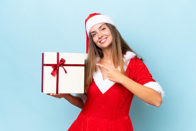 Jeune femme caucasienne habillée en maman noel tenant un cadeau isolé sur fond bleu et le pointant