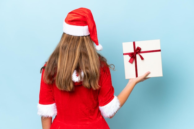 Jeune femme caucasienne habillée en maman noel tenant un cadeau isolé sur fond bleu en position arrière