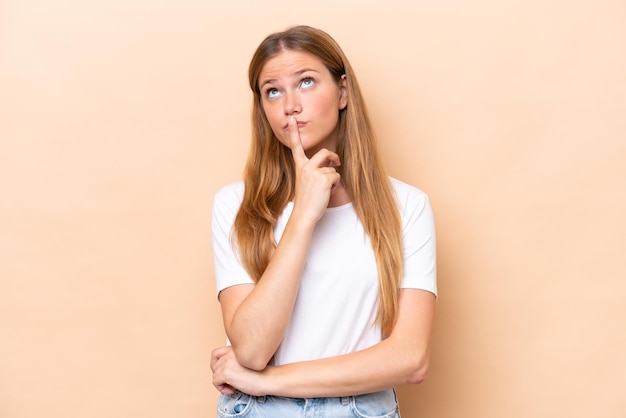 Jeune femme caucasienne isolée sur fond beige ayant des doutes en levant les yeux