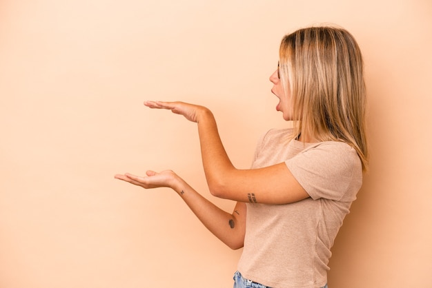 Jeune femme caucasienne isolée sur fond beige choquée et étonnée tenant un espace de copie entre les mains.