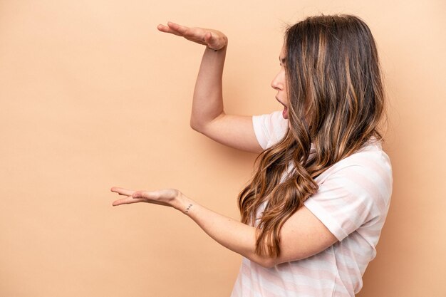 Jeune femme caucasienne isolée sur fond beige choquée et étonnée tenant un espace de copie entre les mains