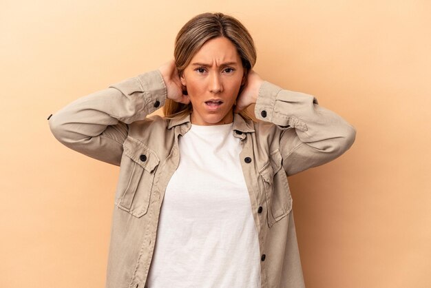 Jeune femme caucasienne isolée sur fond beige couvrant les oreilles avec les mains essayant de ne pas entendre un son trop fort.