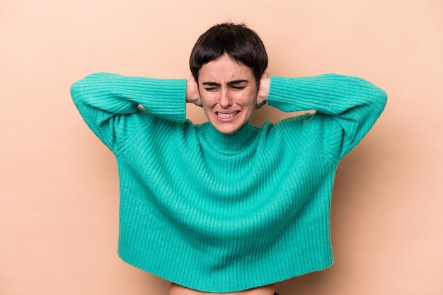 Jeune femme caucasienne isolée sur fond beige couvrant les oreilles avec les mains.