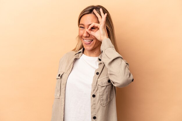 Jeune femme caucasienne isolée sur fond beige excitée en gardant un geste ok sur les yeux.