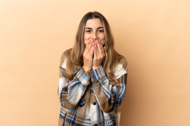 Jeune femme caucasienne isolée sur fond beige heureuse et souriante couvrant la bouche avec les mains