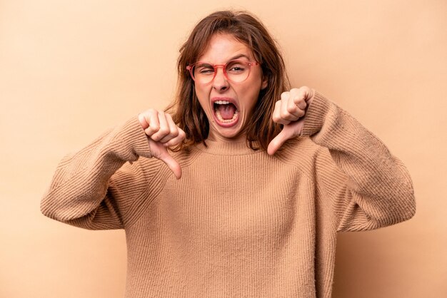 Jeune femme caucasienne isolée sur fond beige montrant le pouce vers le bas et exprimant son aversion