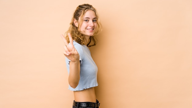 Jeune femme caucasienne isolée sur fond beige montrant le signe de la victoire et souriant largement.