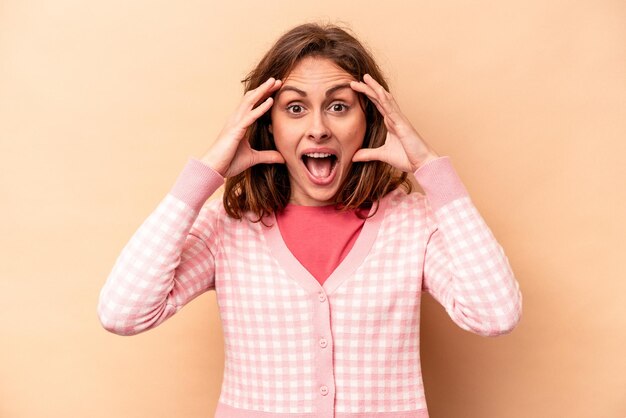 Jeune femme caucasienne isolée sur fond beige recevant une agréable surprise excitée et levant les mains