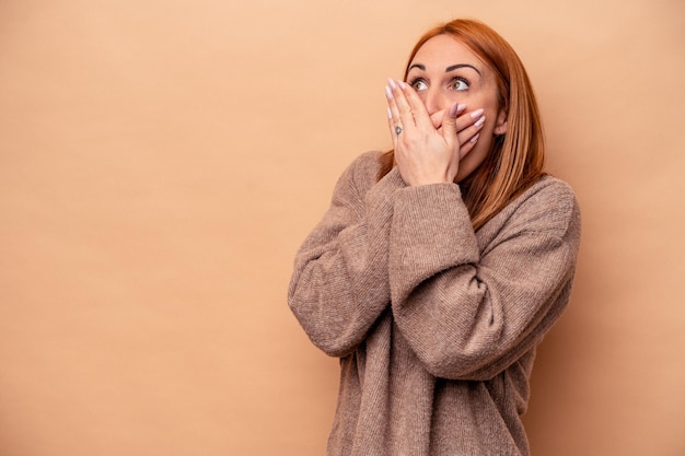 Jeune femme caucasienne isolée sur fond beige réfléchie regardant un espace de copie couvrant la bouche avec la main.