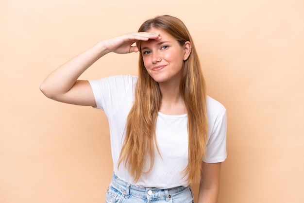 Jeune femme caucasienne isolée sur fond beige regardant loin avec la main pour regarder quelque chose