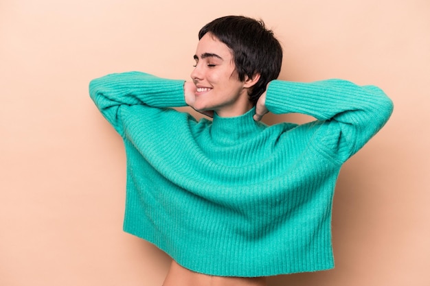 Jeune femme caucasienne isolée sur fond beige se sentant confiante, les mains derrière la tête.