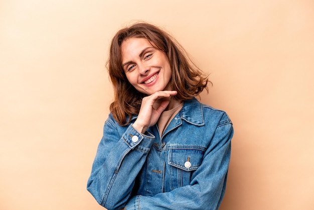 Jeune femme caucasienne isolée sur fond beige souriant heureux et confiant touchant le menton avec la main