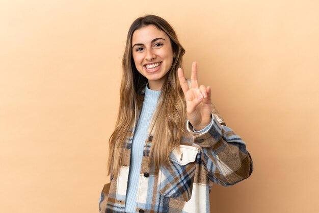 Jeune femme caucasienne isolée sur fond beige souriant et montrant le signe de la victoire