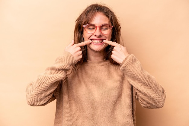 Jeune femme caucasienne isolée sur fond beige sourit pointant du doigt la bouche