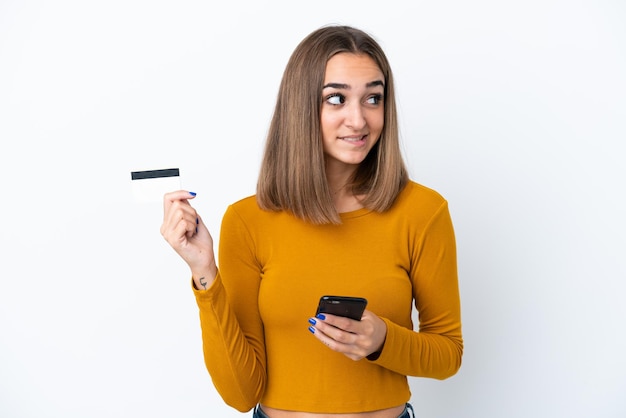 Jeune femme caucasienne isolée sur fond blanc achetant avec le mobile avec une carte de crédit en pensant