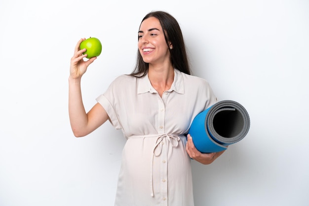 Jeune femme caucasienne isolée sur fond blanc enceinte tenant une pomme et aller à des cours de yoga