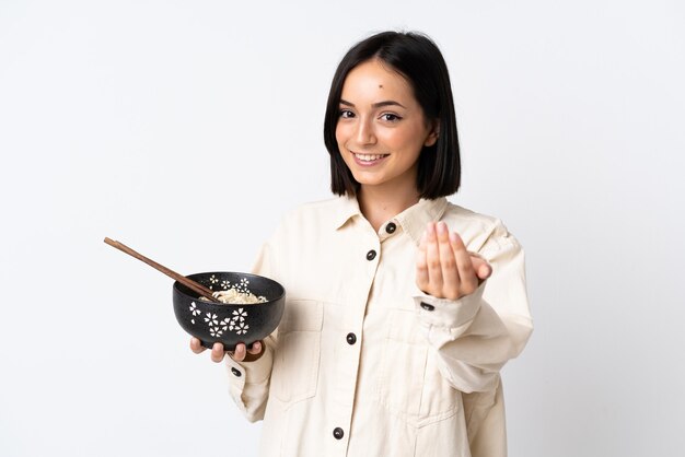 Jeune femme caucasienne isolée sur fond blanc invitant à venir avec la main. Heureux que tu sois venu en tenant un bol de nouilles avec des baguettes