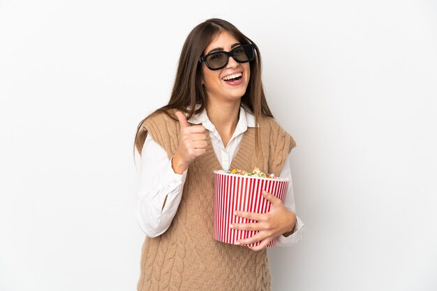 Jeune femme caucasienne isolée sur fond blanc avec des lunettes 3d et tenant un grand seau de pop-corn tout en pointant vers l'avant