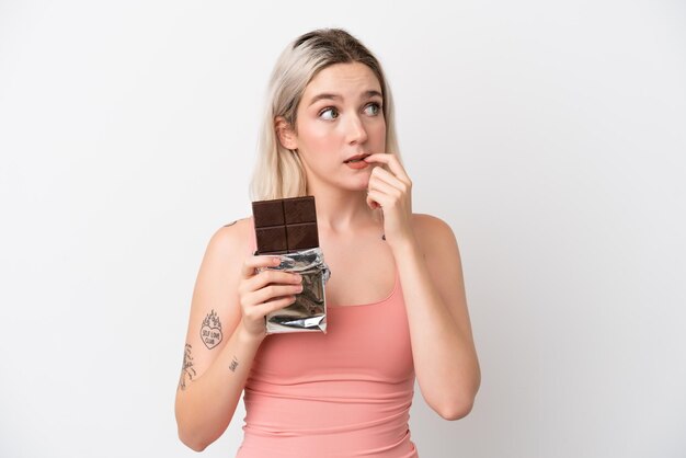 Jeune femme caucasienne isolée sur fond blanc prenant une tablette de chocolat et ayant des doutes