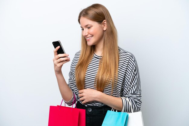 Jeune femme caucasienne isolée sur fond blanc tenant des sacs à provisions et écrivant un message avec son téléphone portable à un ami