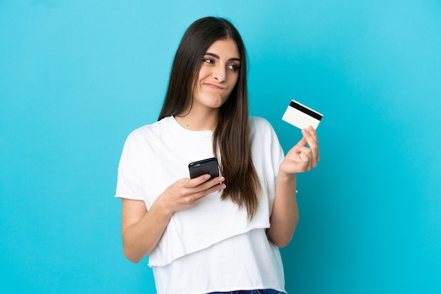 Jeune femme caucasienne isolée sur fond bleu achetant avec le mobile avec une carte de crédit en pensant