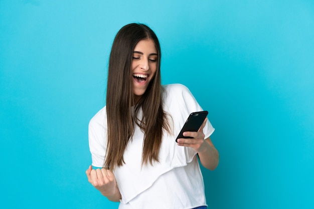 Jeune femme caucasienne isolée sur fond bleu à l'aide d'un téléphone portable et faisant un geste de victoire