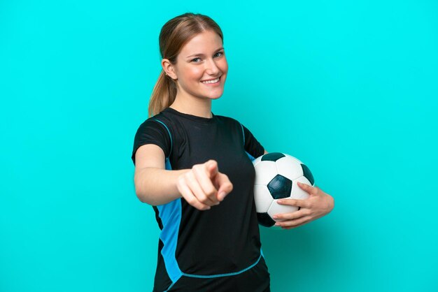 Jeune femme caucasienne isolée sur fond bleu avec ballon de football et pointant vers l'avant