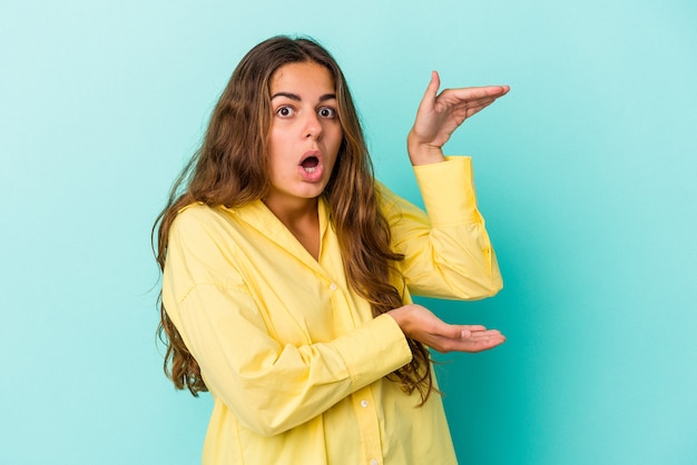 Jeune femme caucasienne isolée sur fond bleu choquée et étonnée tenant un espace de copie entre les mains.