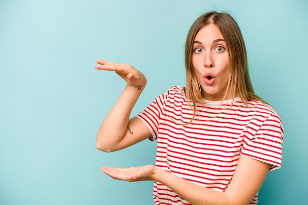 Jeune femme caucasienne isolée sur fond bleu choquée et étonnée tenant un espace de copie entre les mains