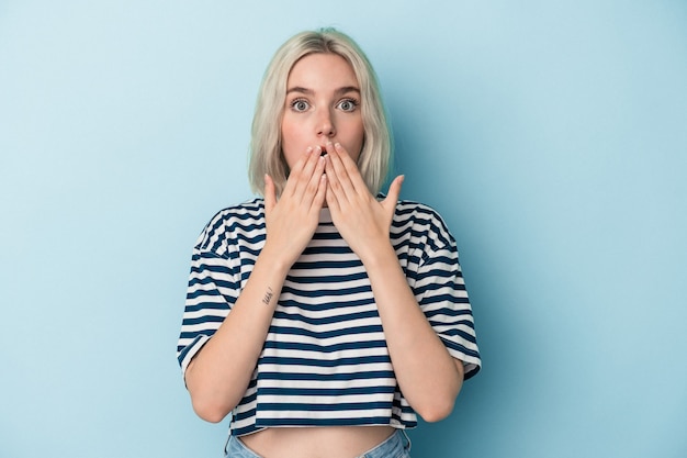 Jeune femme caucasienne isolée sur fond bleu choquée, se couvrant la bouche avec les mains, impatiente de découvrir quelque chose de nouveau.