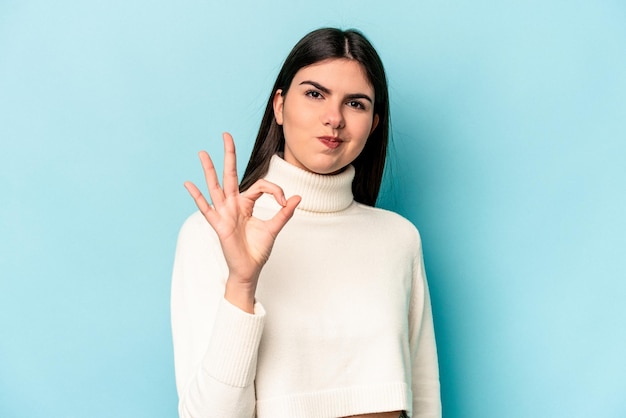 Jeune femme caucasienne isolée sur fond bleu cligne de l'œil et tient un geste correct avec la main