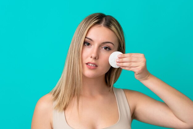 Jeune femme caucasienne isolée sur fond bleu avec un coton pour enlever le maquillage de son visage