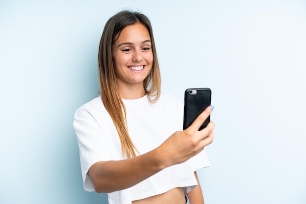 Jeune femme caucasienne isolée sur fond bleu faisant un selfie avec téléphone portable