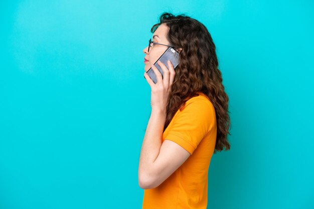 Jeune femme caucasienne isolée sur fond bleu gardant une conversation avec le téléphone portable avec quelqu'un