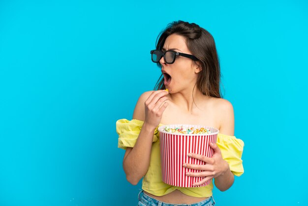 Jeune femme caucasienne isolée sur fond bleu avec des lunettes 3d et tenant un grand seau de pop-corn tout en regardant de côté