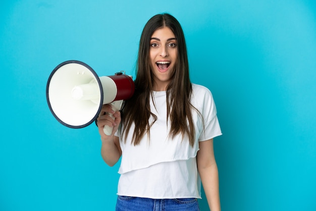 Jeune femme caucasienne isolée sur fond bleu tenant un mégaphone et avec une expression de surprise