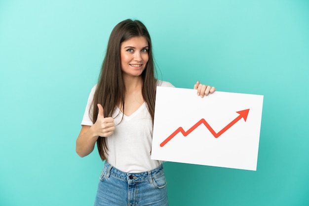 Jeune femme caucasienne isolée sur fond bleu tenant une pancarte avec un symbole de flèche de statistiques croissantes avec le pouce vers le haut