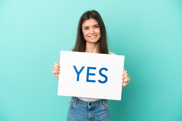 Jeune femme caucasienne isolée sur fond bleu tenant une pancarte avec texte OUI avec une expression heureuse