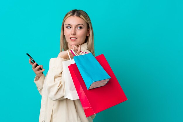 Jeune femme caucasienne isolée sur fond bleu tenant des sacs à provisions et un téléphone portable