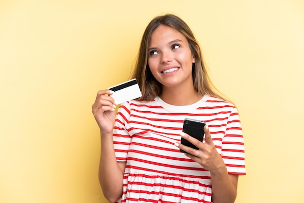 Jeune femme caucasienne isolée sur fond jaune achetant avec le mobile avec une carte de crédit en pensant