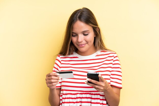 Jeune femme caucasienne isolée sur fond jaune achetant avec le mobile avec une carte de crédit