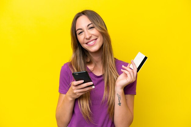 Jeune femme caucasienne isolée sur fond jaune achetant avec le mobile avec une carte de crédit