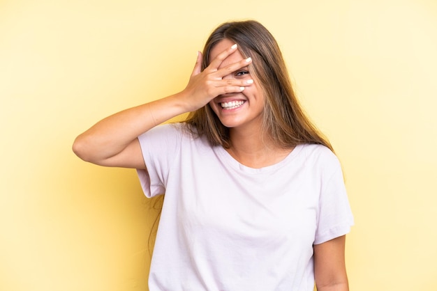 Jeune femme caucasienne isolée sur fond jaune couvrant les yeux par les mains et souriant