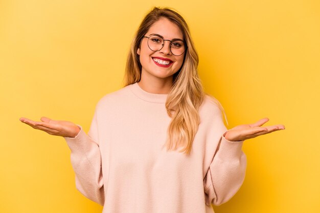 Jeune femme caucasienne isolée sur fond jaune fait de l'échelle avec les bras se sent heureuse et confiante